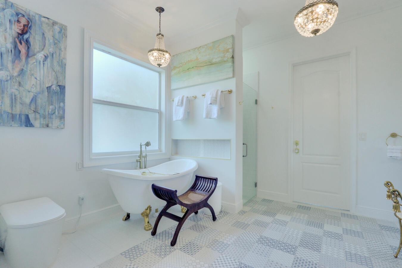 Opulent white master bathroom featuring a vintage clawfoot soaking tub with gold feet, geometric mosaic tile flooring, a hanging crystal chandelier, and fine art.