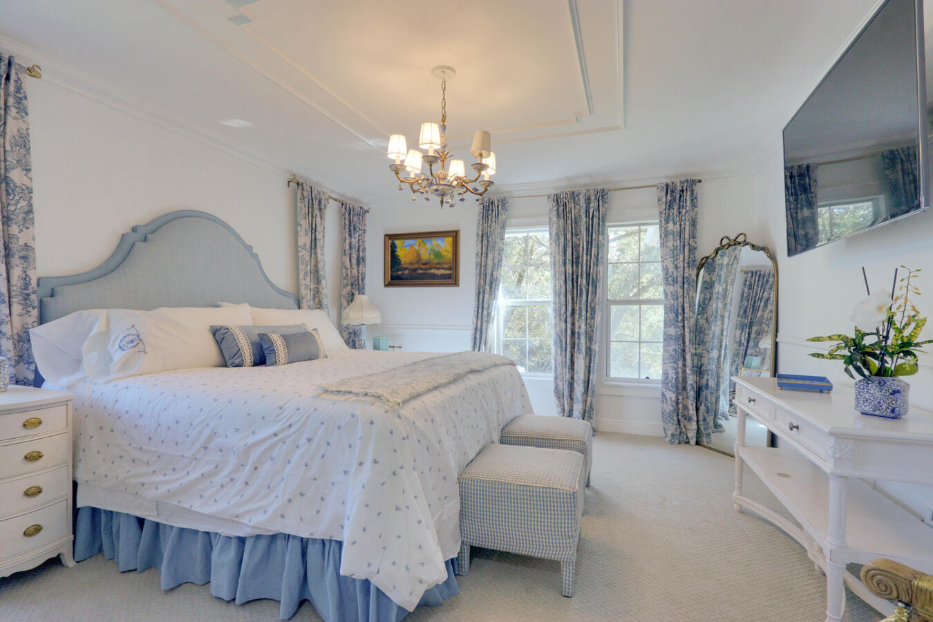 Romantic French-style bedroom with a blue upholstered headboard, matching blue toile de Jouy curtains and bedding, an ornate gold full-length mirror, and a crystal chandelier.