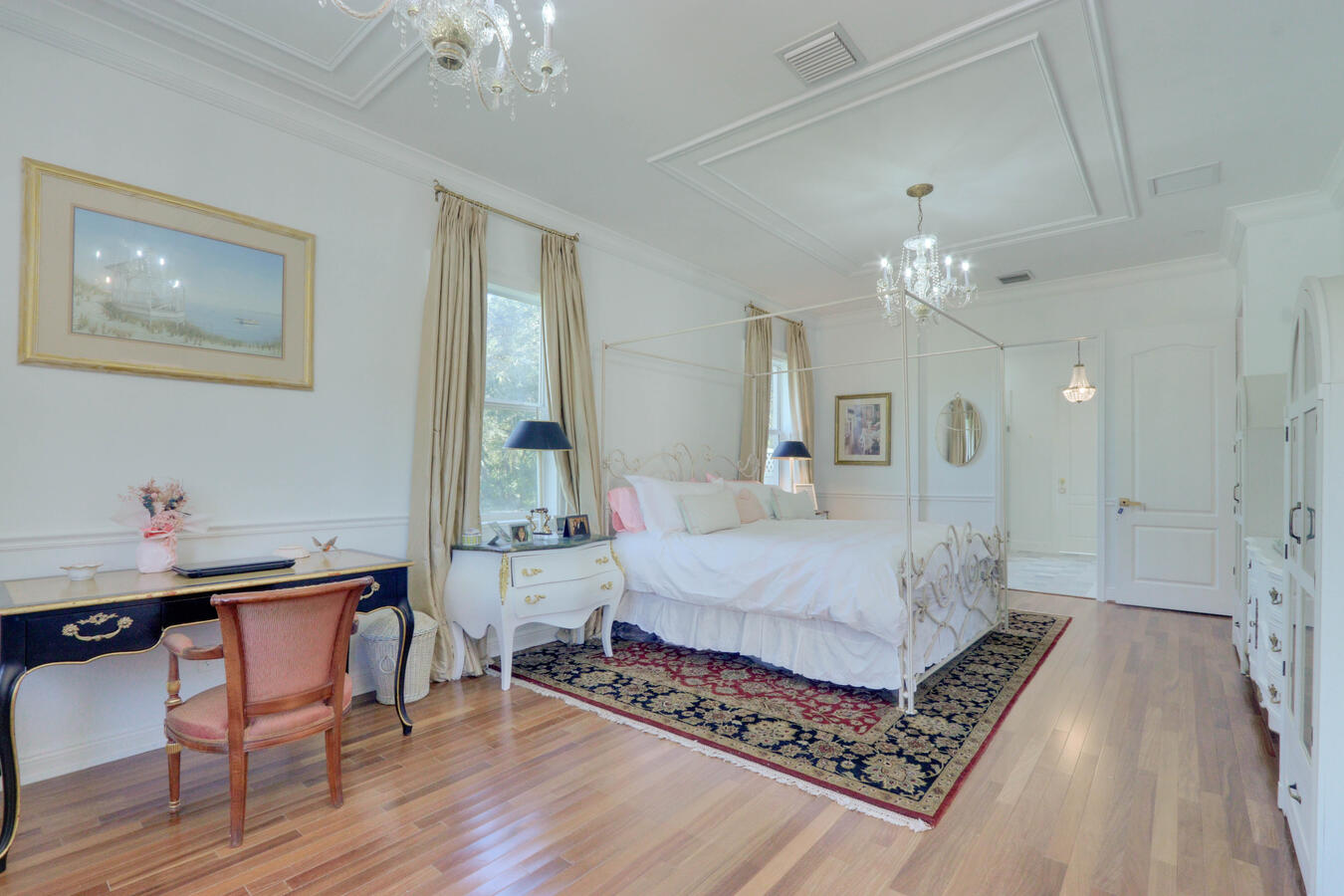 Bright luxury master bedroom featuring a white iron four-poster canopy bed, dark ornate oriental rug over hardwood floors, and a crystal chandelier.