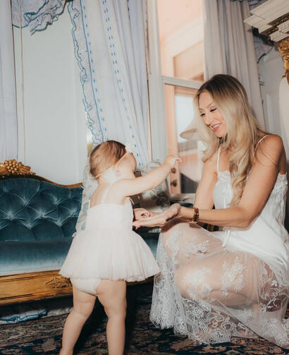 A mother dressed in a white silk slip shares a tender moment holding hands with her standing baby girl on a vintage rug in front of a blue velvet and gold sofa.