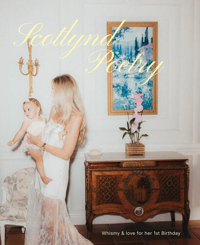 Editorial portrait of a mother holding her baby daughter next to a vintage French marquetry commode chest and a gold-framed painting in a sun-drenched luxury studio venue.