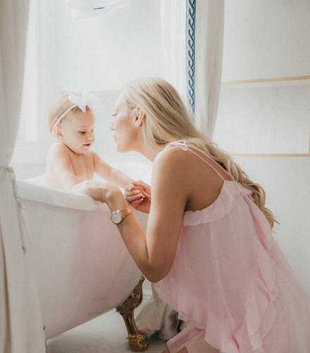 A mother in a soft pink ruffled dress kneels next to a vintage white clawfoot bathtub with gold legs, holding hands with her baby girl during a bright, airy family portrait session at Sarti Luxe Estates.