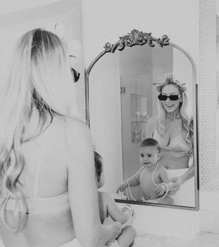 Black and white high-fashion editorial photograph of a mother with hair rollers holding her infant daughter in front of dramatic silk window drapes at a luxury film location.