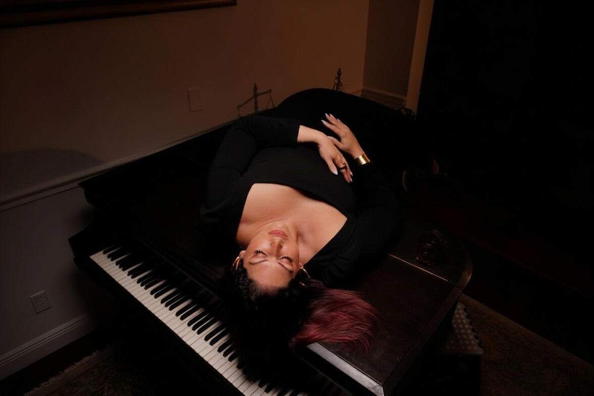Luxury editorial portrait of a woman in a sequined black cape sitting on a classic black grand piano under dramatic warm lighting.